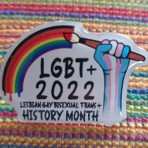 The badge design shows a hand grasping a paintbrush with a pride rainbow flowing from the tip. The hand is coloured in with the blue, white and pink, the colours of the trans pride flag. The text on the badge reads Lesbian Gay Bisexual Trans plus History Month
